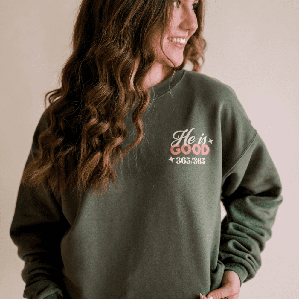 Woman wearing forest green sweatshirt with front chest print that says "He is Good 365/365"