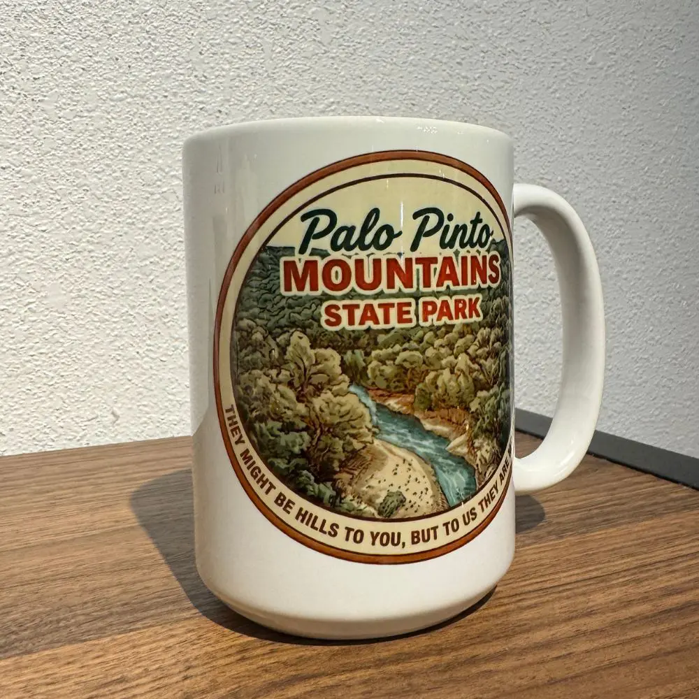 Side angle of ceramic Palo Pinto Mountains State Park mug showing trees, river, and full quote