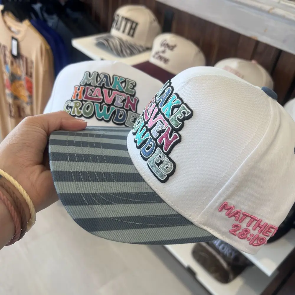 Side view of white Make Heaven Crowded trucker hat featuring colorful embroidery, blue striped bill, and Matthew 28:19 embroidered on the side.