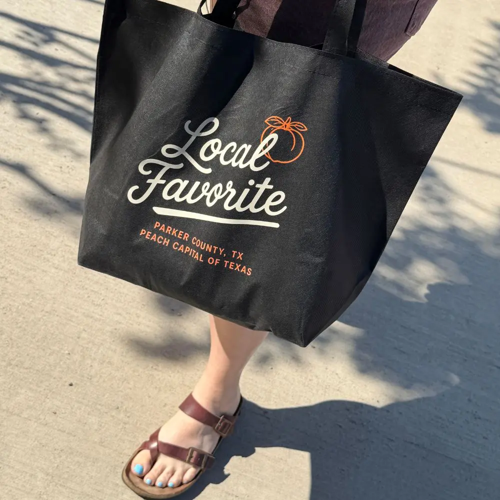 Person carrying oversized black Parker County Peach Festival tote bag featuring Local Favorite peach design.
