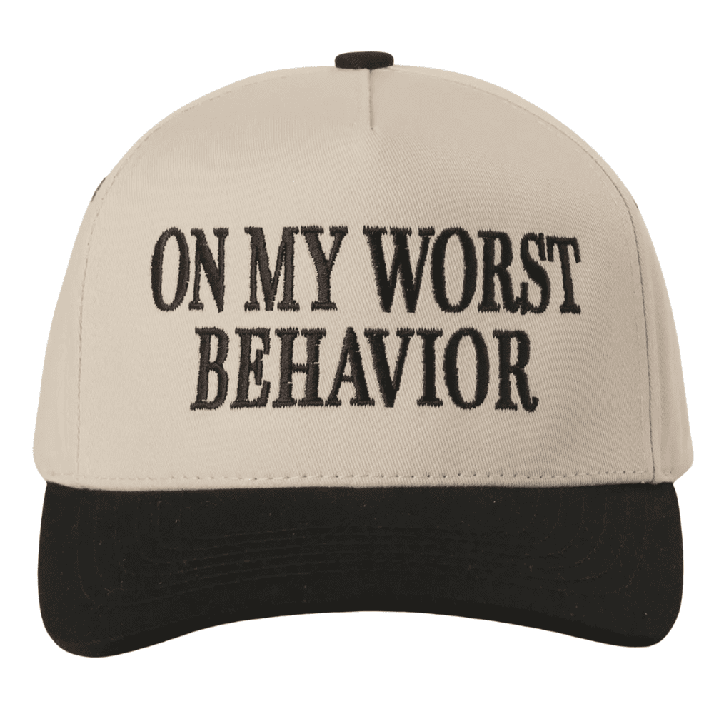 A cream and black trucker hat with bold black embroidery reading “ON MY WORST BEHAVIOR” on the front panel.