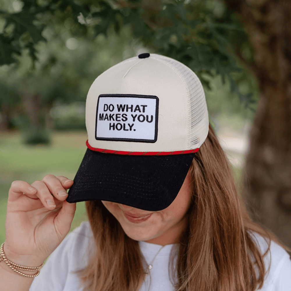 Woman adjusting the back of a cream and black Christian trucker hat that says “Do What Makes You Holy”