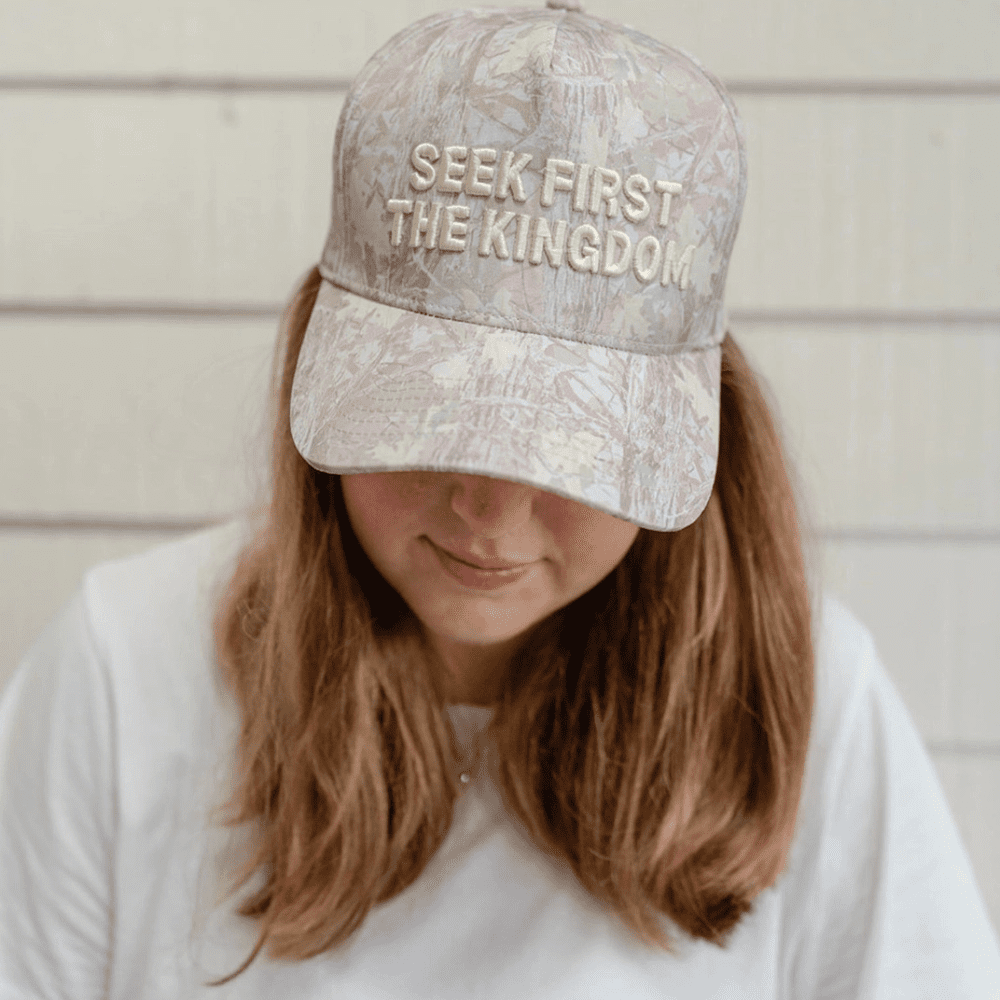 Close-up front view of woman wearing a camouflage-pattern Seek First The Kingdom hat