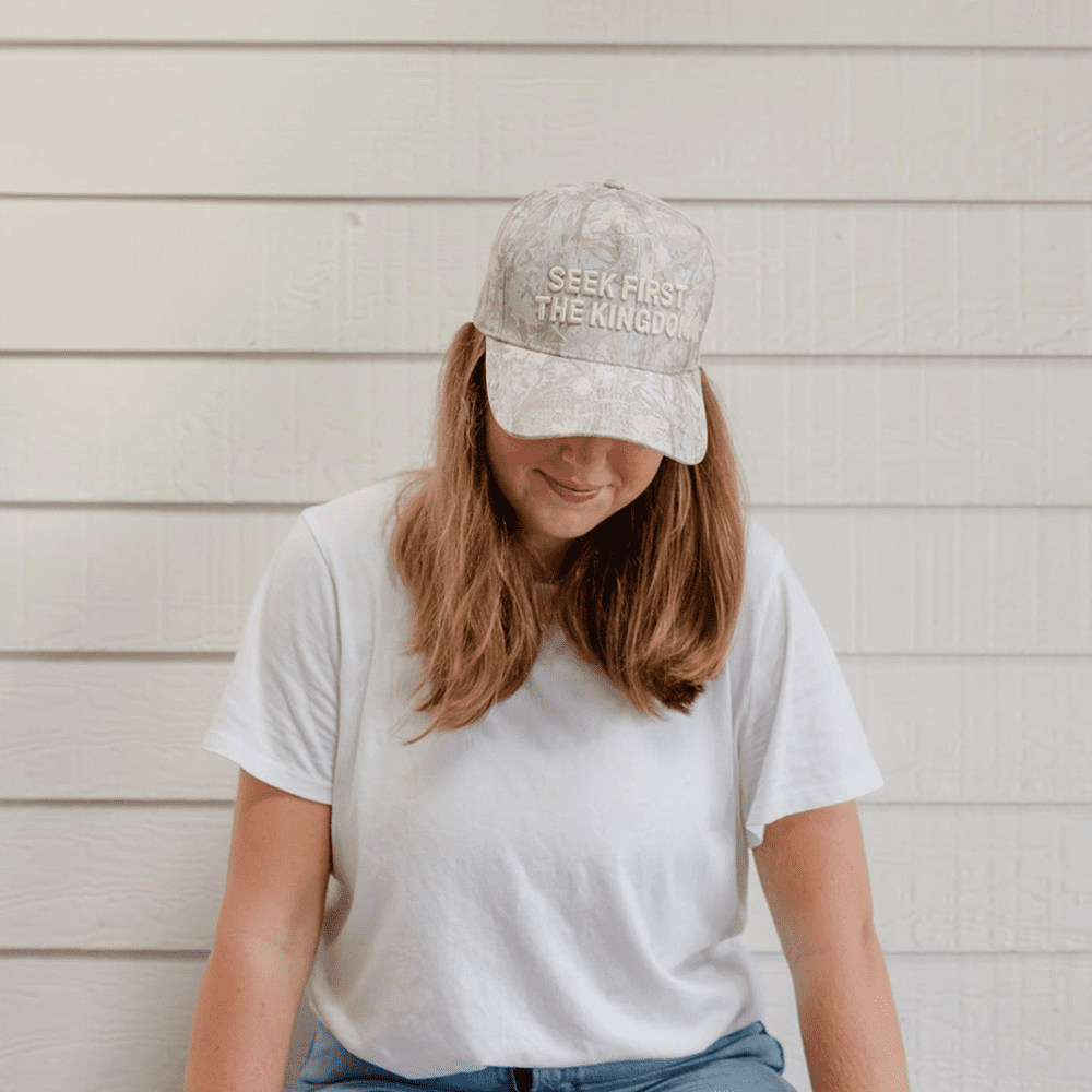Woman sitting against white siding wearing Seek First The Kingdom camo hat